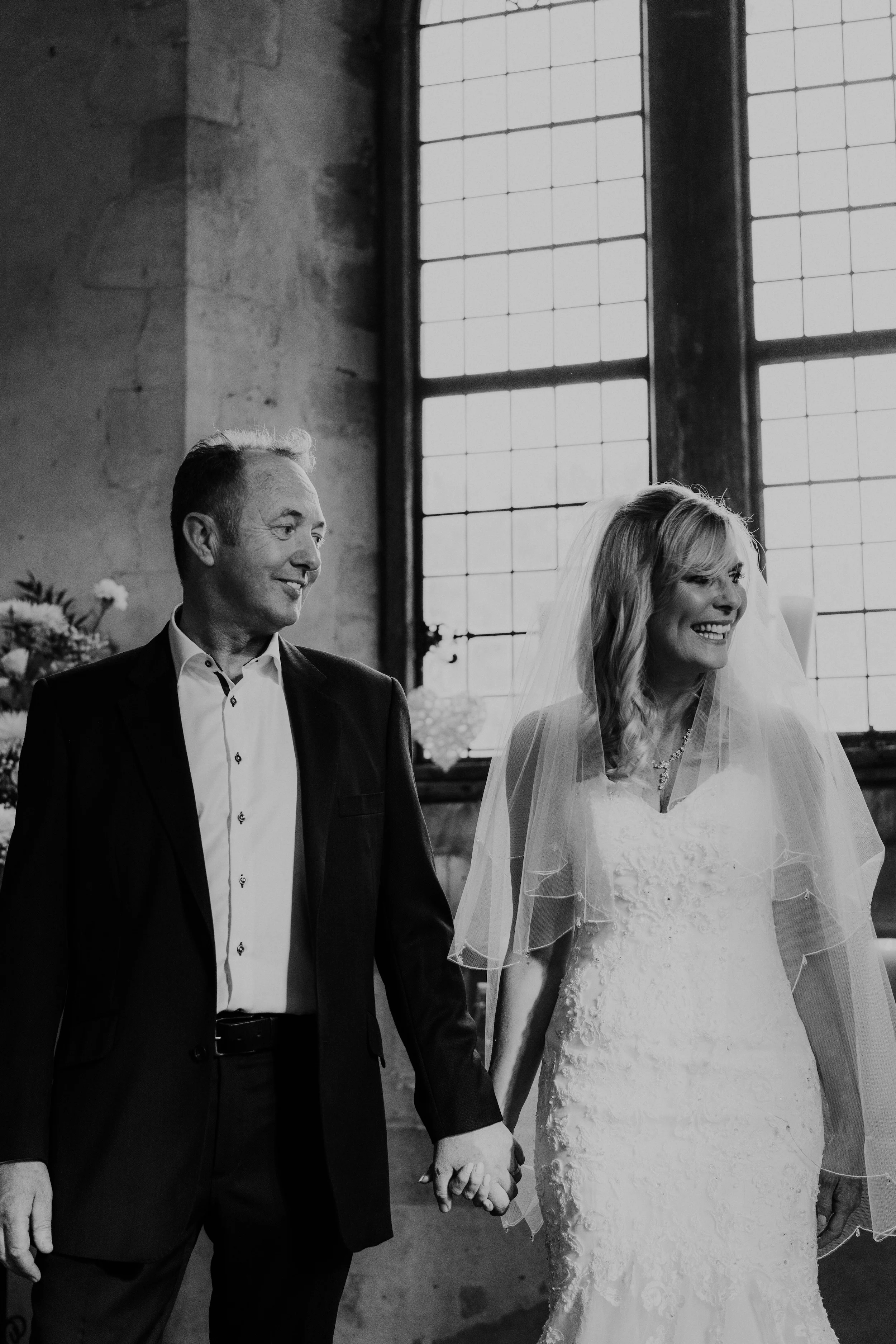 Black and white photograph of Mark and Fiona walking hand in hand past a tall priory window