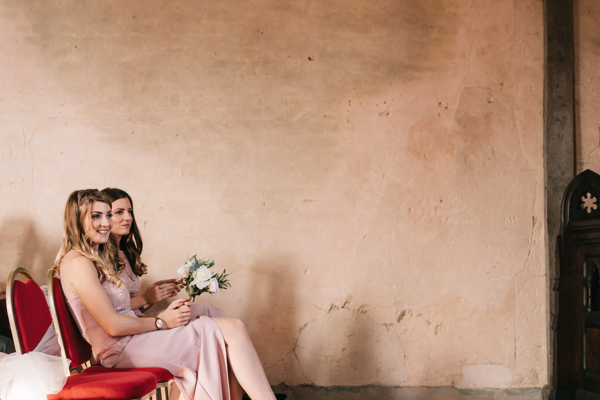 Two bridesmaids sit together against the old plaster wall of the Great Hall
