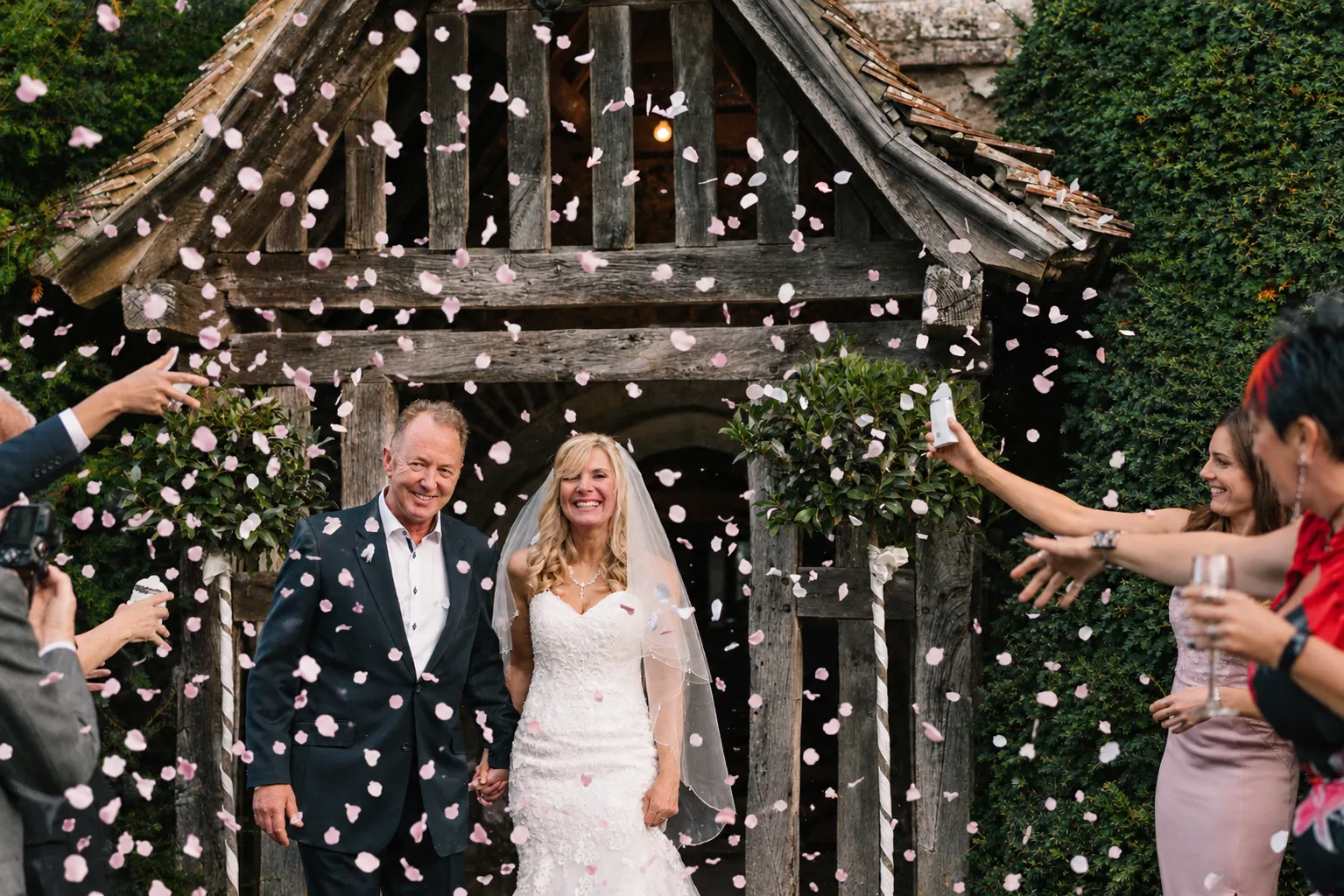 Pink petal confetti fills the air around Mark and Fiona at the priory porch