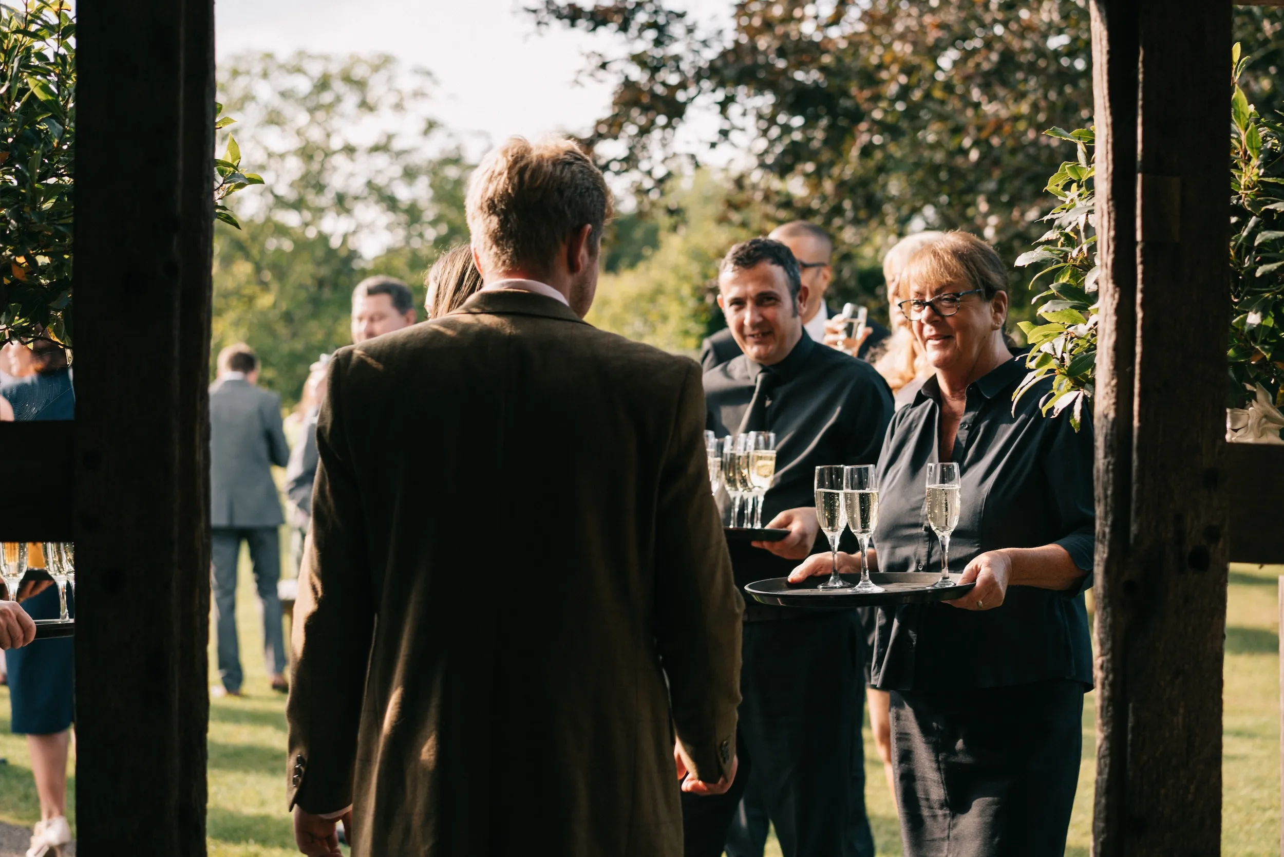 Staff carry trays of champagne out to guests on the lawn, framed by timber posts