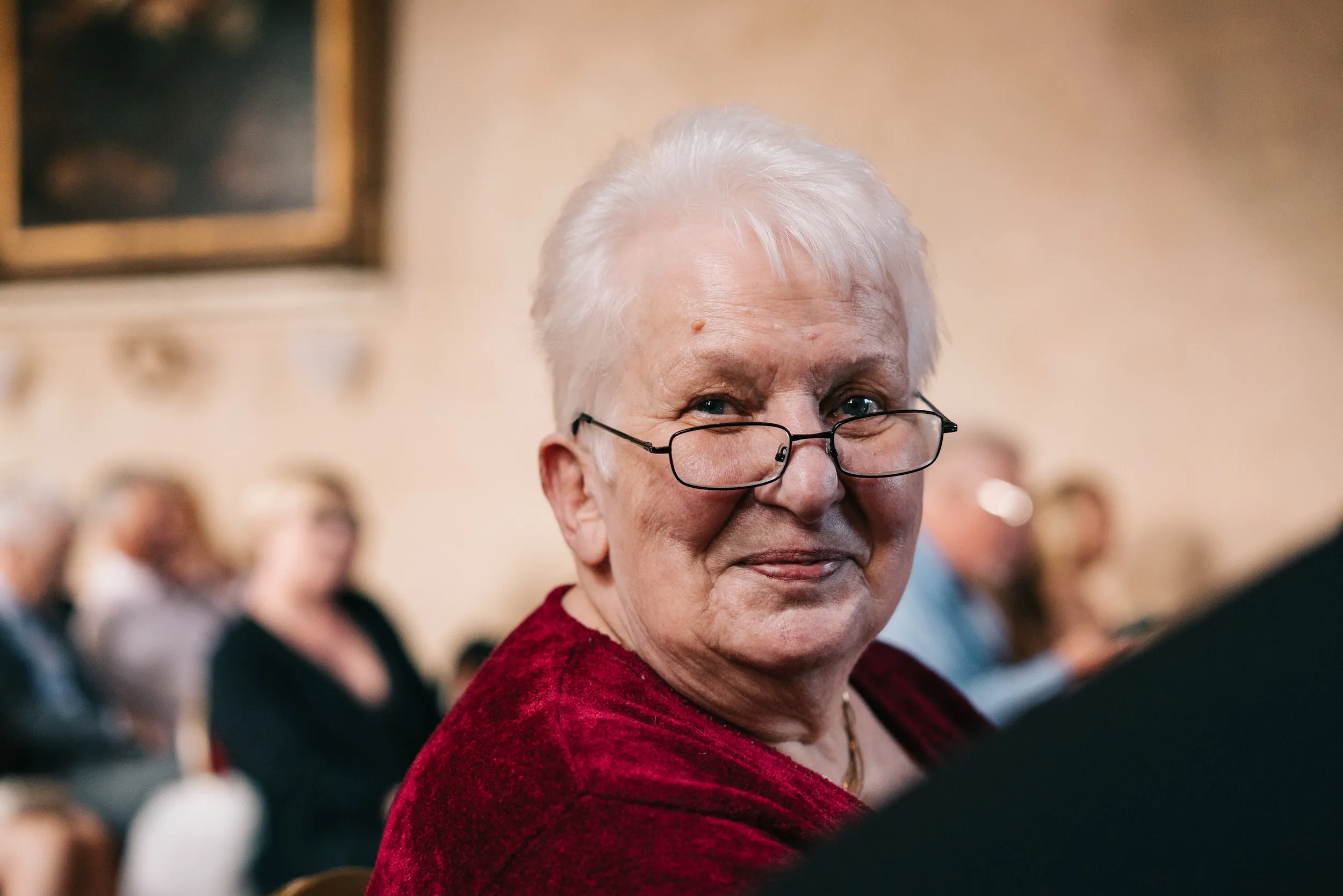 An older guest in red velvet turns to smile during the ceremony