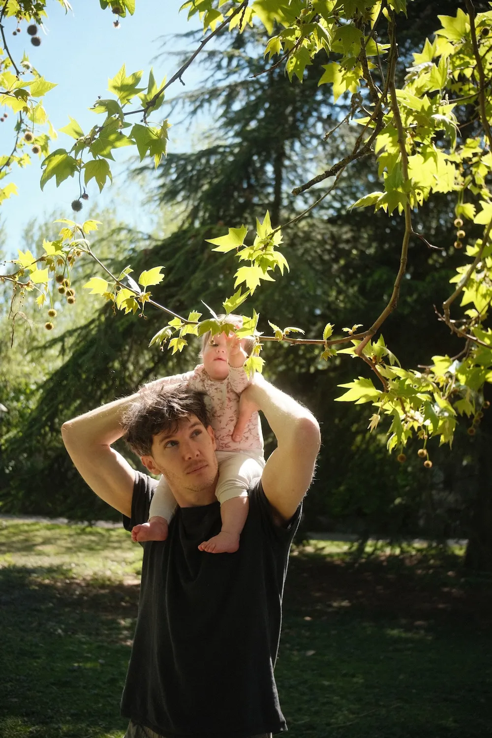 A man carries a baby on his shoulders under a plane tree in the grounds of The Friars