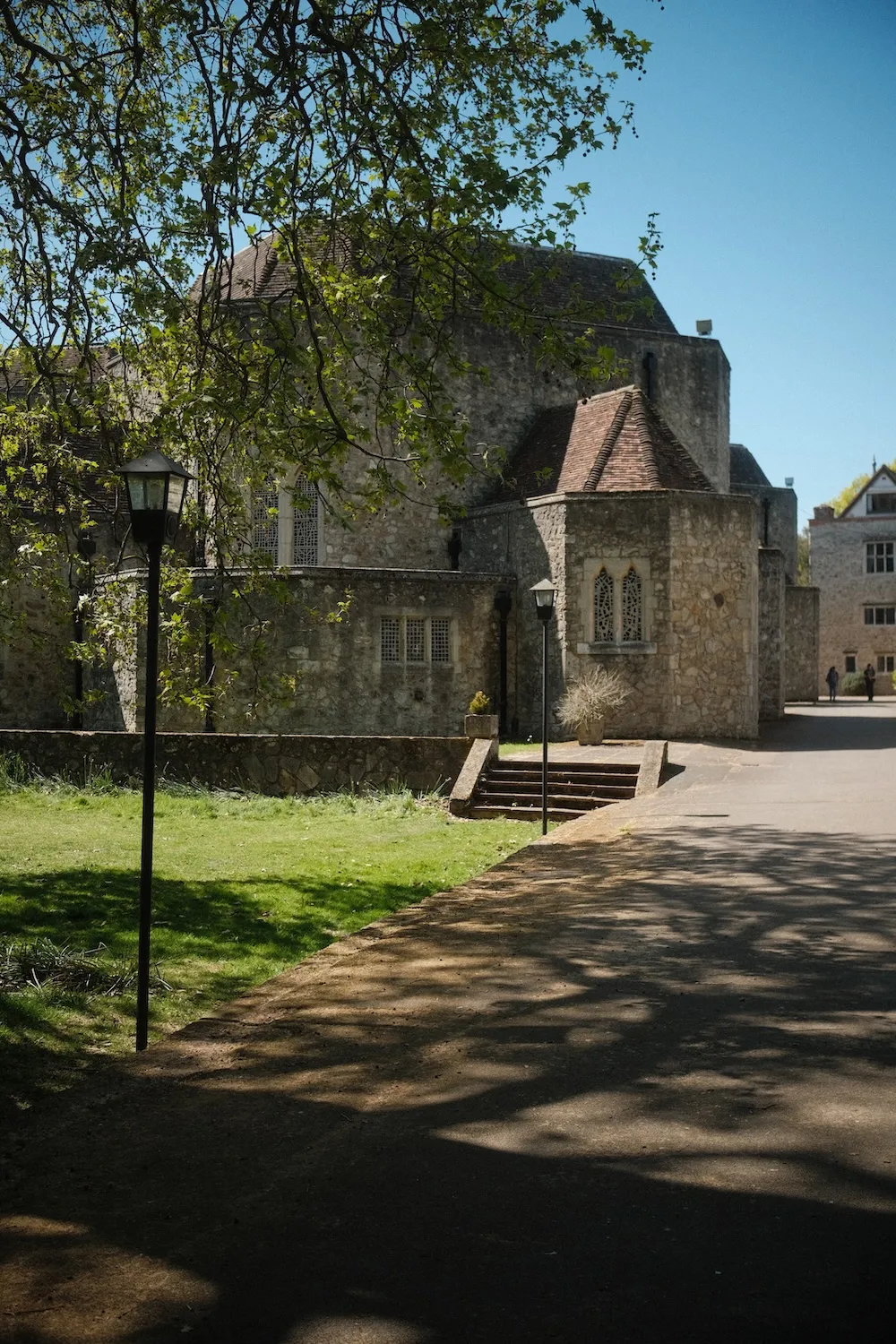 A shaded path through the grounds of The Friars, with the old stone priory buildings beyond